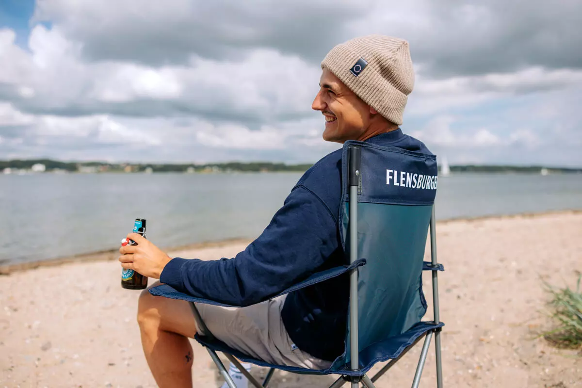 Junger Mann sitzt auf einem Strandstuhl am Strand und trinkt ein Flensburger Strand-Lager.