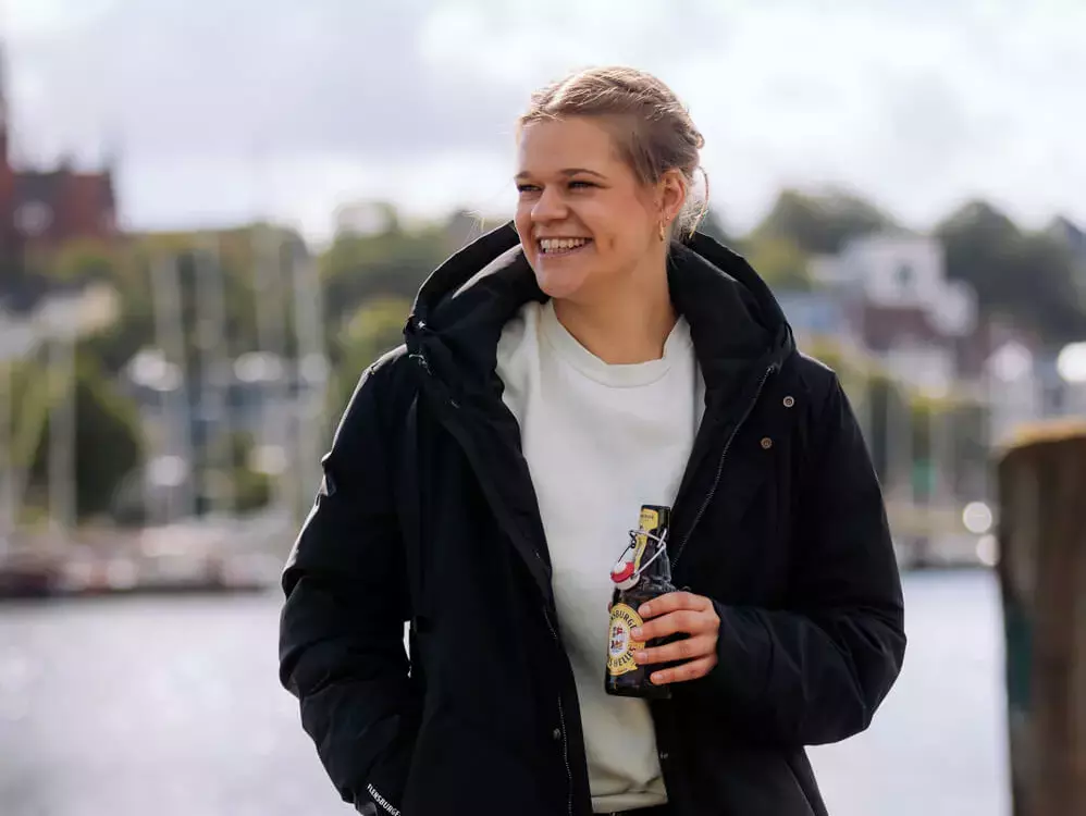 Eine junge Frau mit einem dunklen Mantel steht mit einem FLENS in der Hand am Hafen.