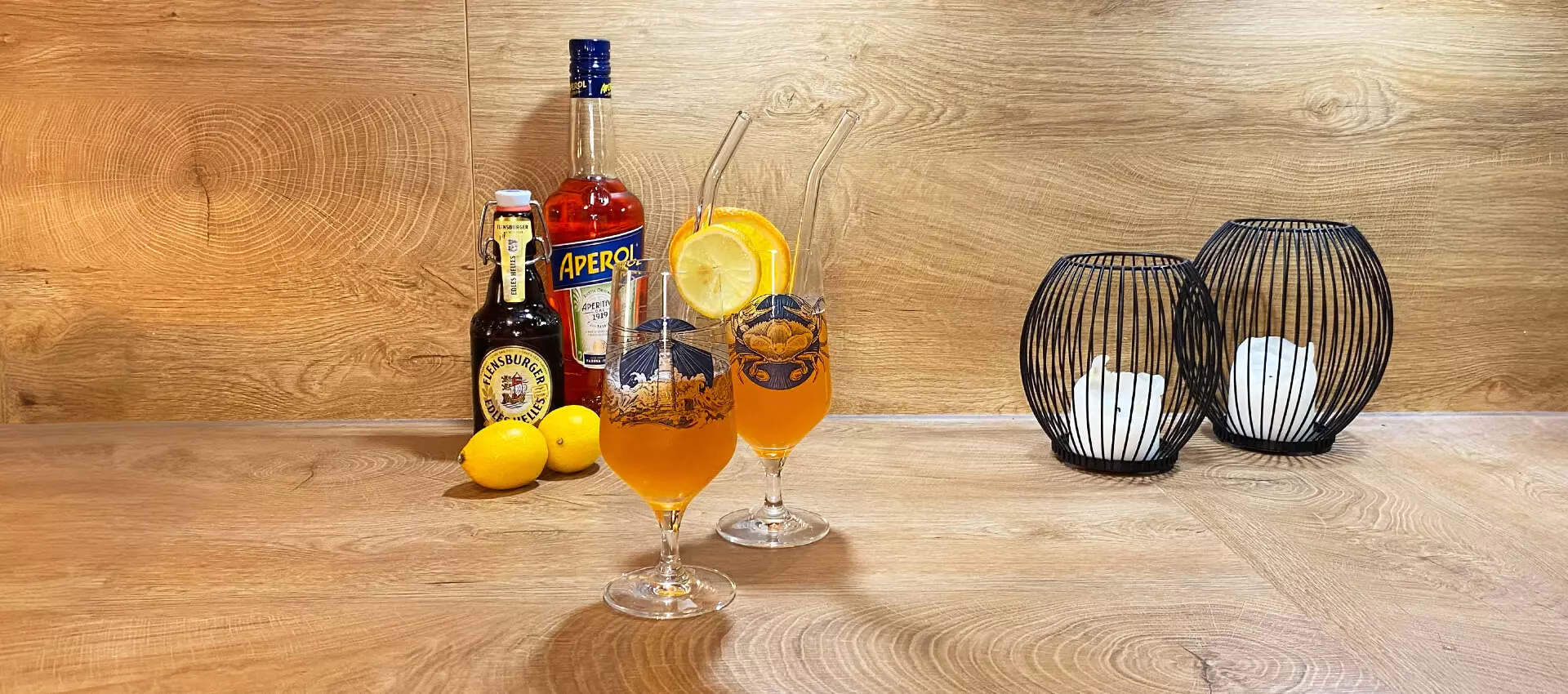 Zwei Gläser mit Aperol-Bier-Cocktail auf Holzuntergrund, im Hintergrund eine Flasche Aperol, Flensburger Edles Helles und zweit Zitronen.