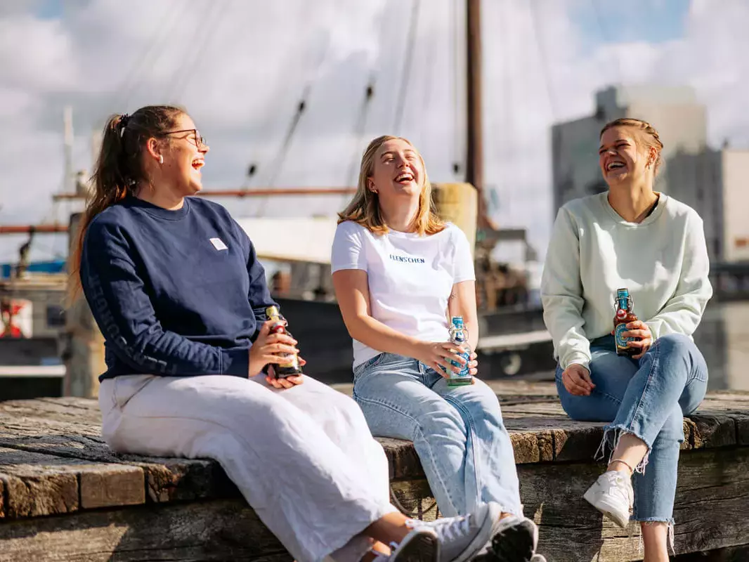 Drei junge Frauen sitzen am Hafen, trinken FLENS und lachen.