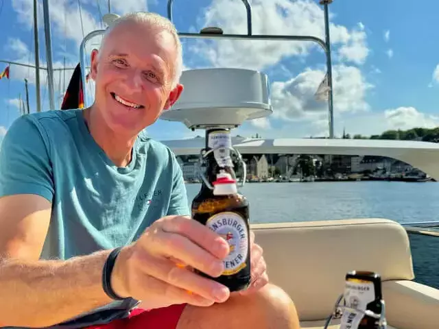 Ein Mann auf einem Segelboot prostet mit einem Flensburger Pilsener.