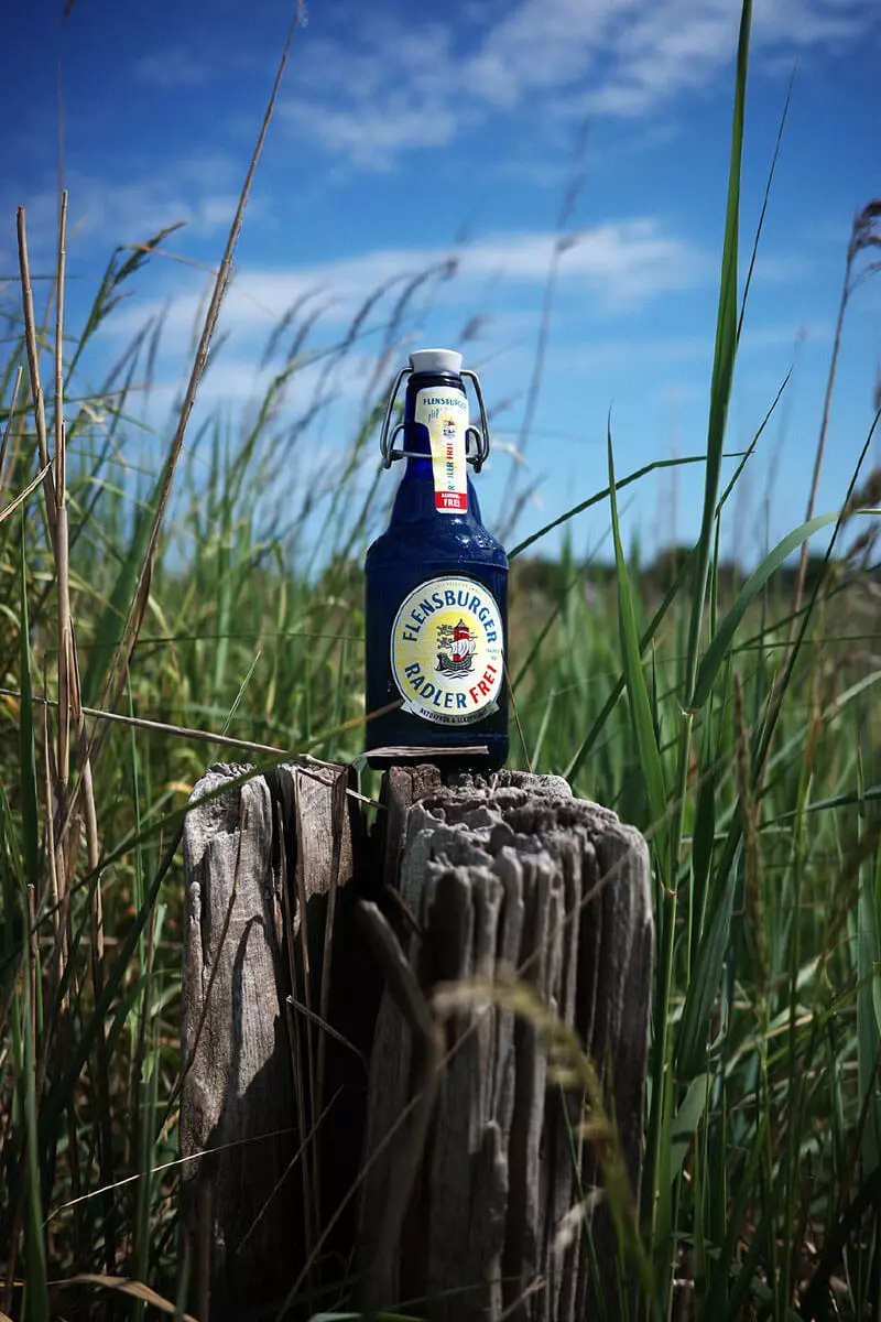 Eine blaue Flasche Flensburger Radler Frei auf einem Holzpoller im Schilfgras.