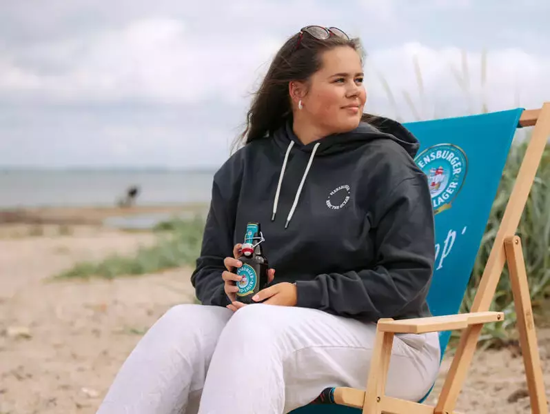 Eine junge Frau sitzt in einem Strandstuhl am Strand und hält ein Flensburger Strand-Lager in der Hand.