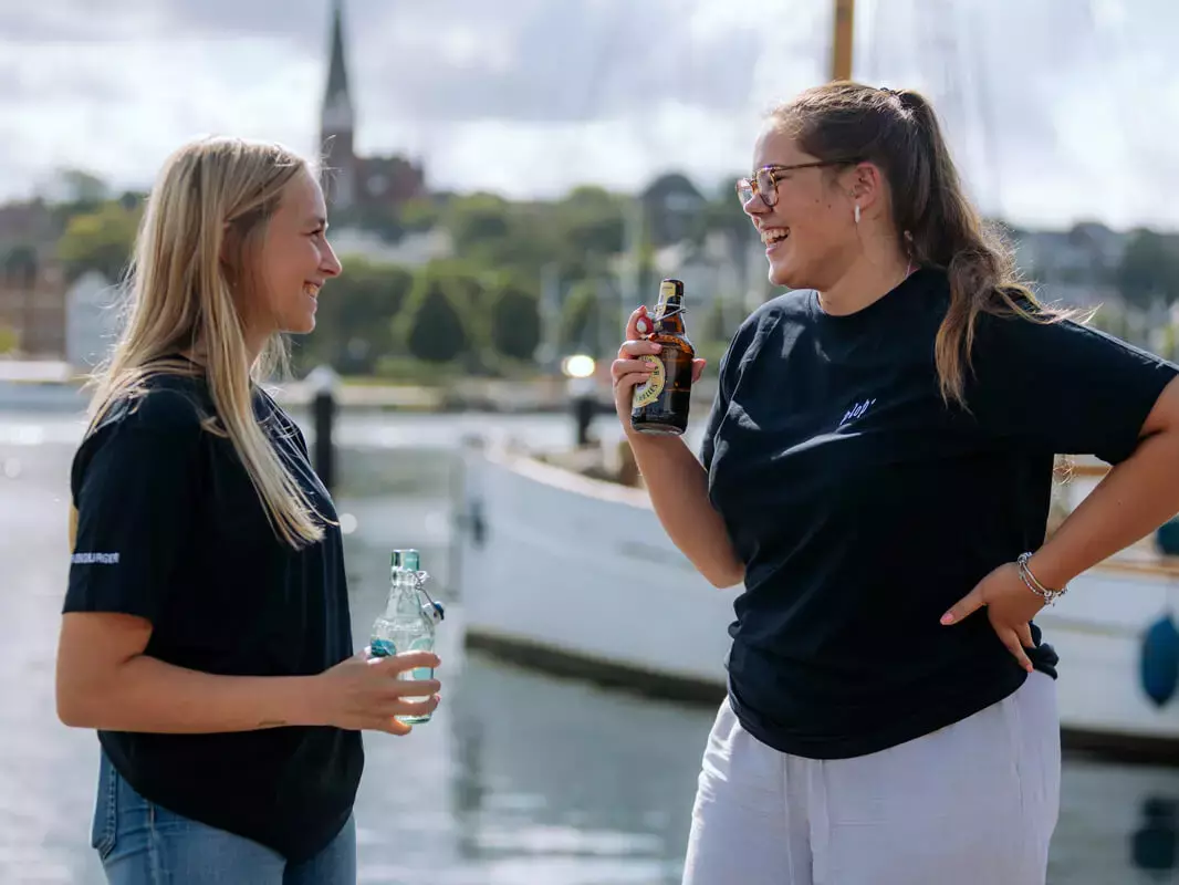 Zwei junge Frauen unterhalten sich am Hafen und trinken FLENS.