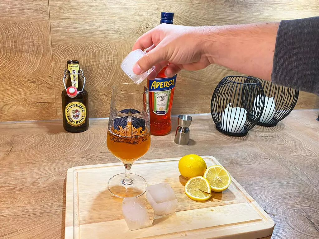 In den Aperol-Bier-Cocktail wird ein Eiswürfel gegeben, daneben Zitronen und im Hintergrund Flensburger Edles Helles.