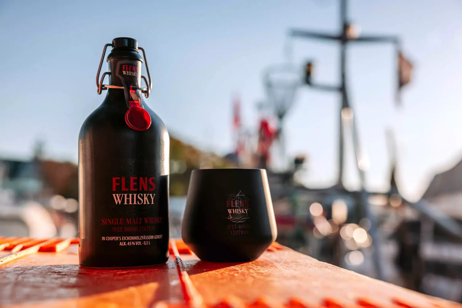 Sylt-Rømø-Edition des FLENS Whiskys in einer schwarzen Tonflasche, daneben das schwarze Whiskyglas, am Hafen.