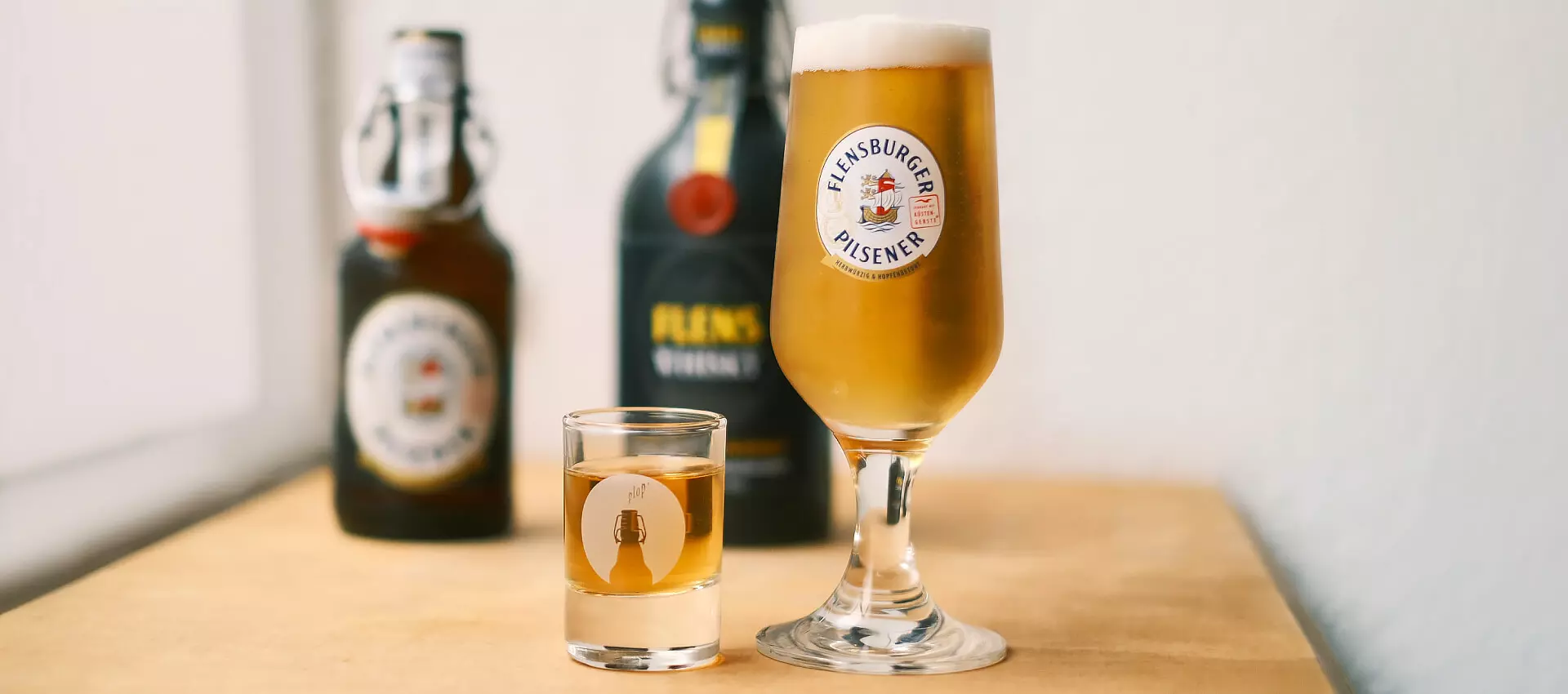 Ein Glas frisch gezapftes Flensburger Pilsener, daneben ein Shotglas mit FLENS Whisky; im Hintergrund etwas verschwommen die dazugehörigen Flaschen.