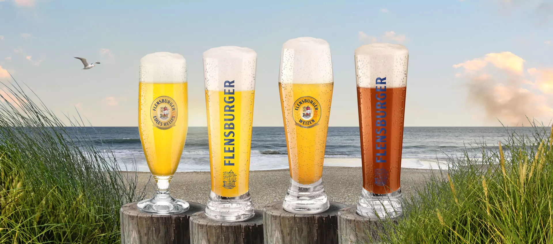 Vier Gläser mit unterschiedlich hellen Bieren auf Pollern am Strand.