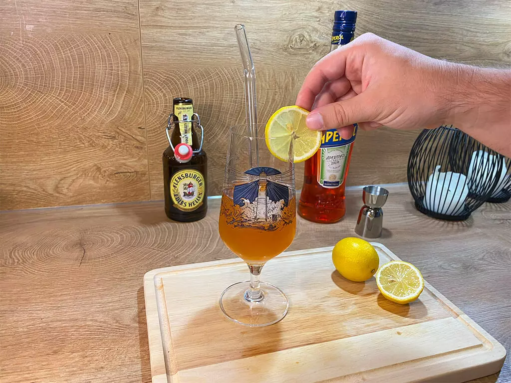 Der Aperol-Bier-Cocktail wird mit einer Zitronenscheibe garniert, im Hintergrund Flensburger Edles Helles.