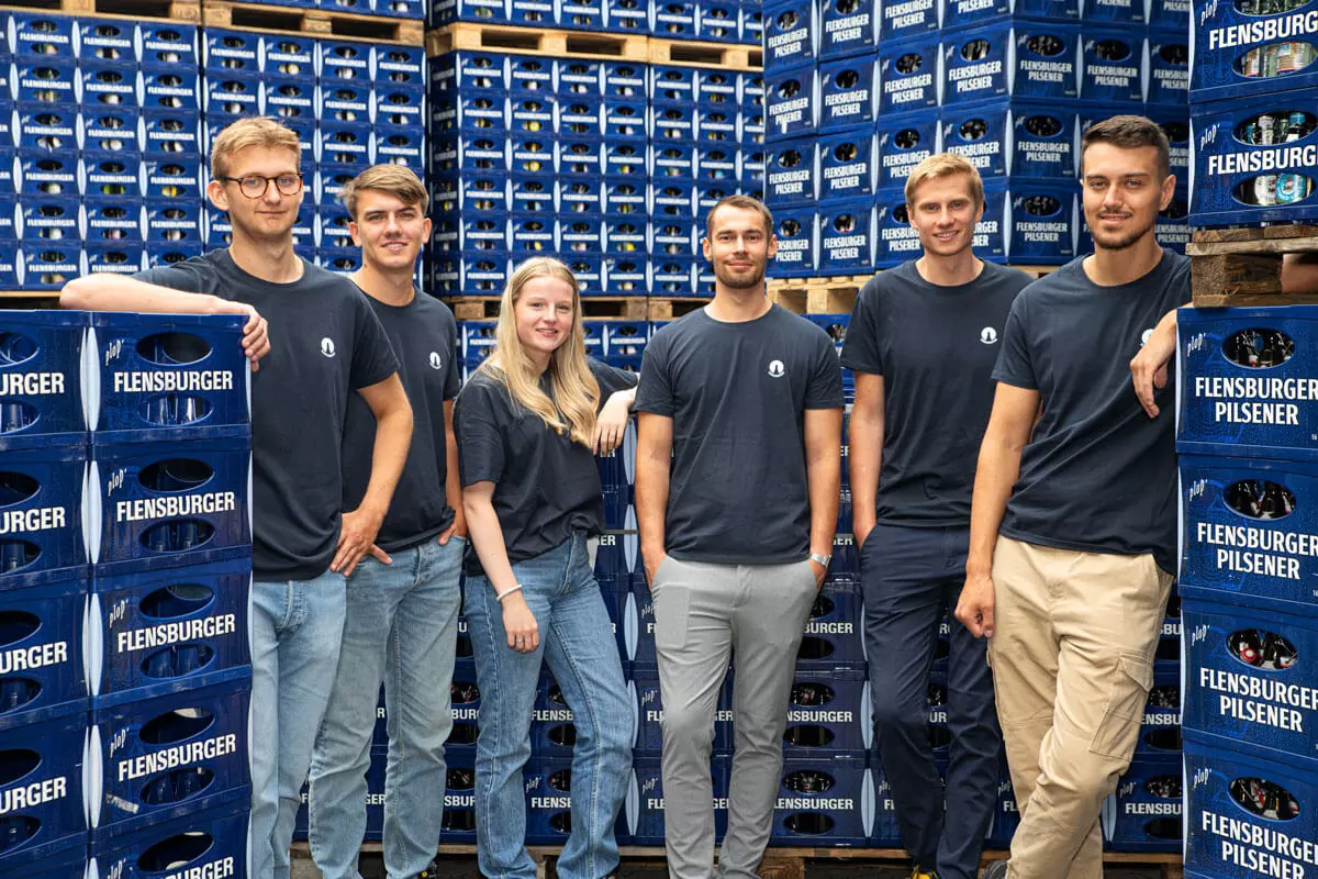 Die sechs neuen Azubis der Flensburger Brauerei aus 2024 inmitten vieler blauer FLENS Kästen.