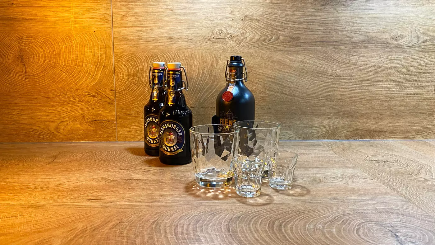 Zwei Flaschen Flensburger Dunkel, eine Flasche FLENS Whisky, zwei Tumbler-Gläser und zwei Shooter-Gläser auf einer Holztheke.