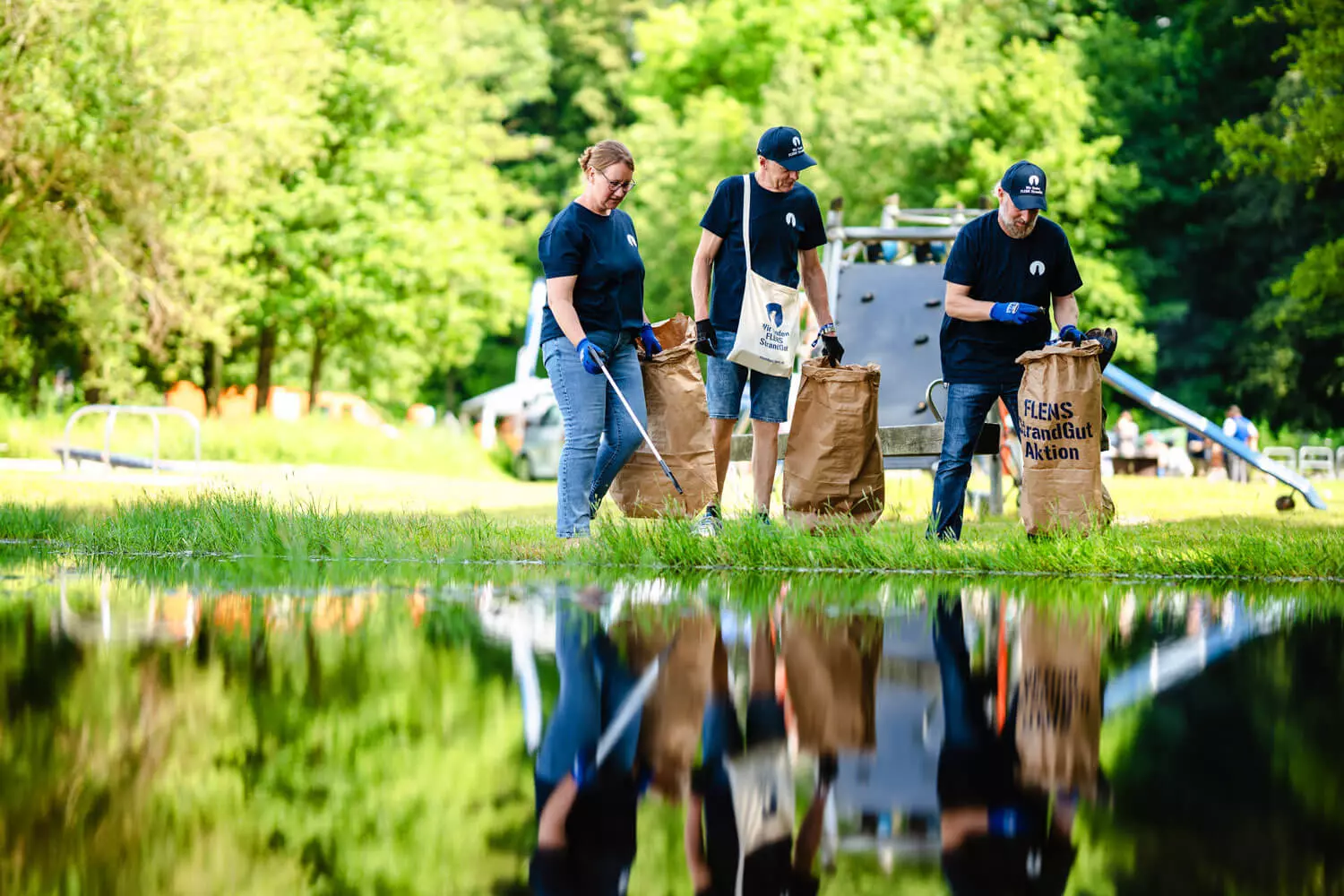 Drei Helfende, die mit Müllsammeltüten und -greifern am Ufer entlanggehen.