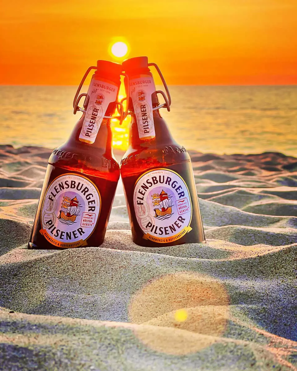 Zwei Flaschen Flensburger Pilsener stehen im Sand aneinander gelehnt und leuchten orange im Sonnenuntergang.