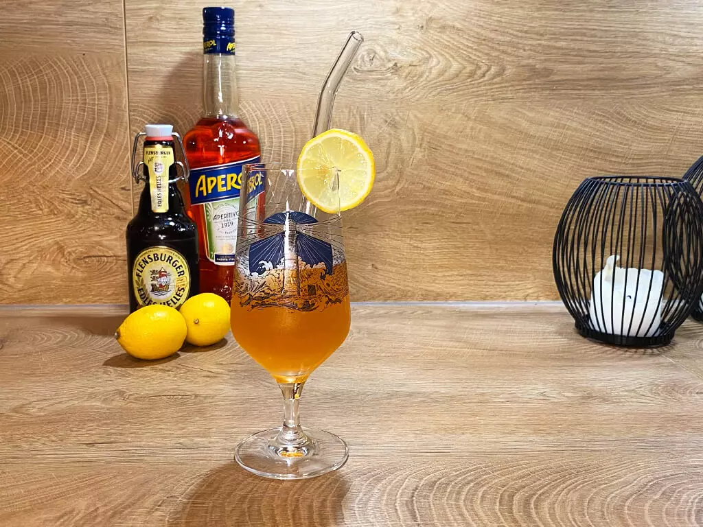 Aperol-Bier-Cocktail im Bierglas, im Hintergrund eine Aperol-Flasche, Zitronen und Flensburger Edles Helles.