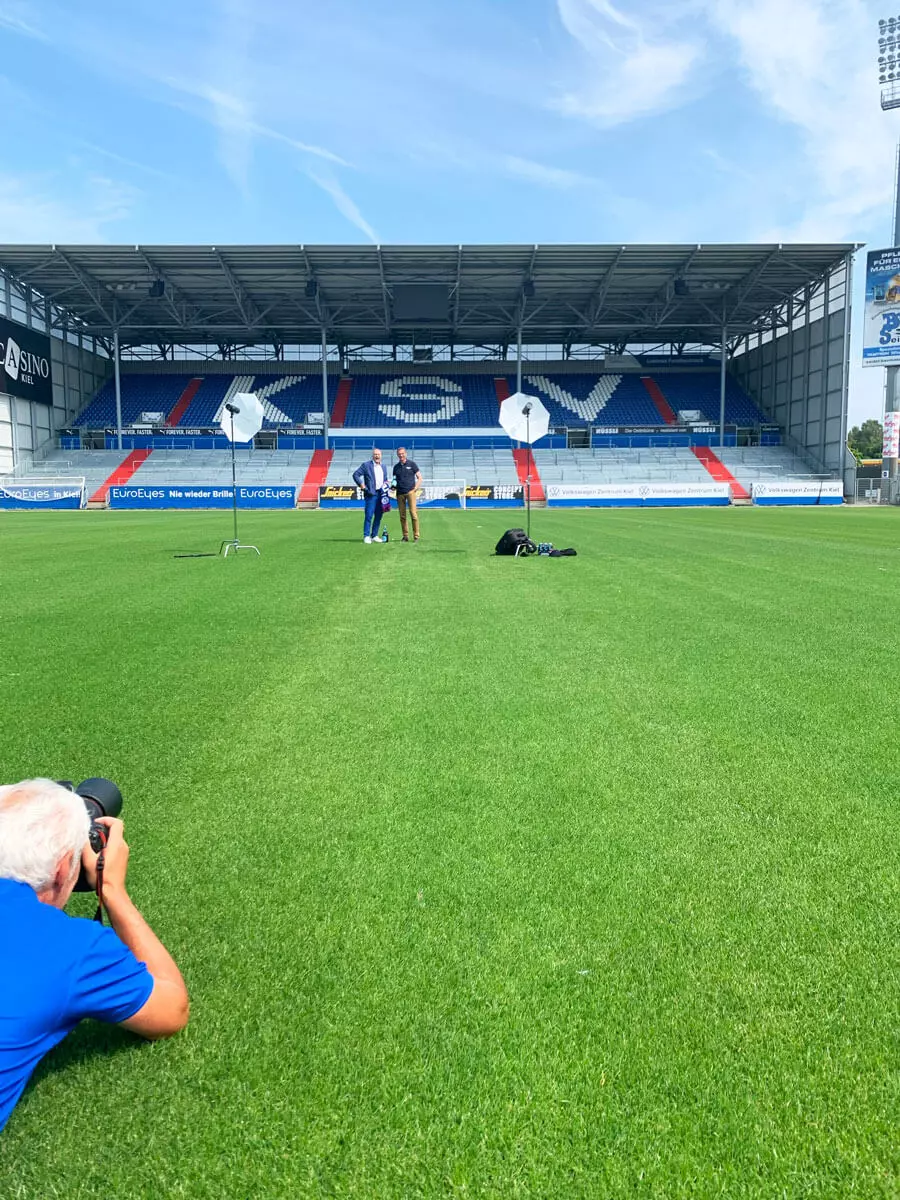 Fotograf liegt auf dem Stadionrasen im Holstein-Stadion und fotografiert die Geschäftsführer vom KSV und FLENS.