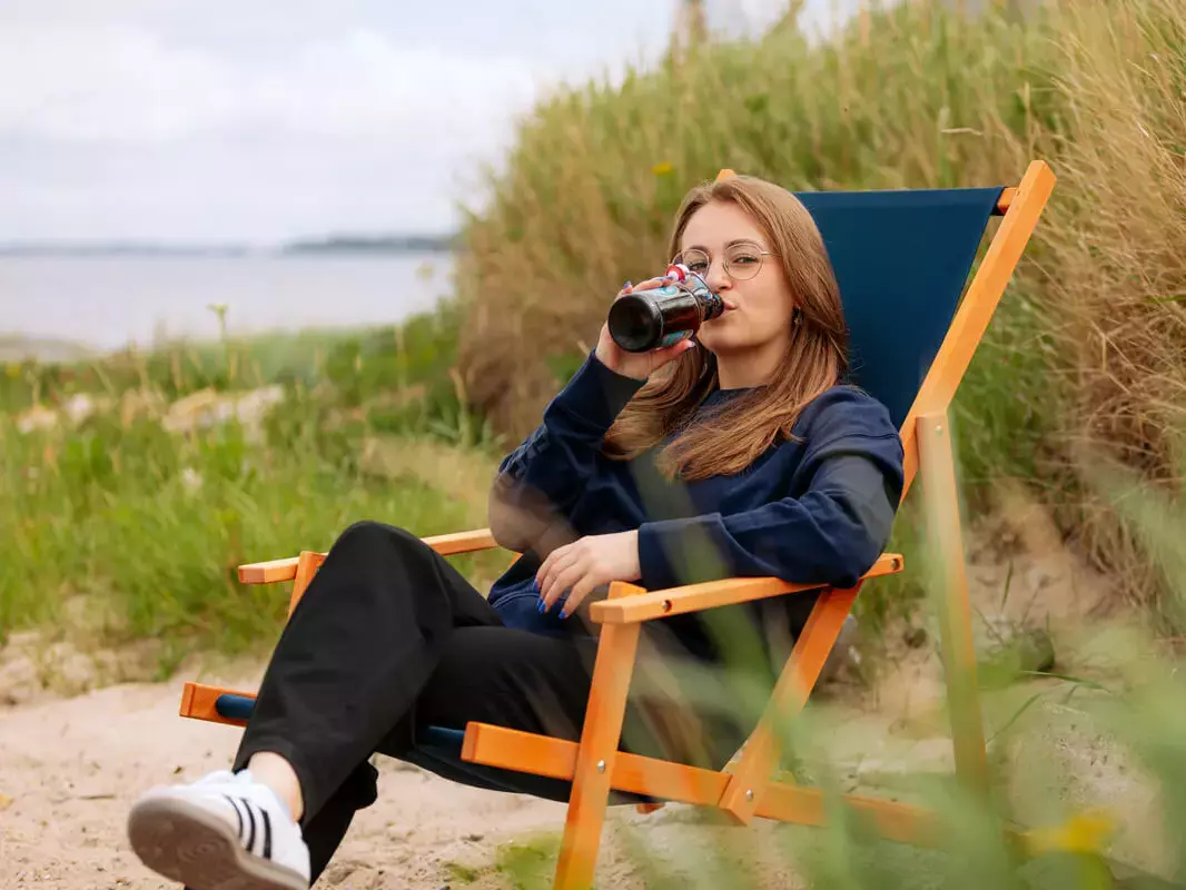 Eine junge Frau sitzt in einem Strandstuhl am Strand und trinkt FLENS.